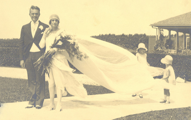 A bride and groom in a photo from the 1920s. The bride's dress has a long train that is held by two small children