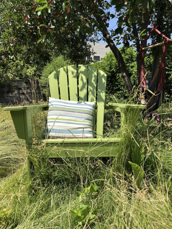 A green Adirondack chair with a pillow, with grass growing in front of it.