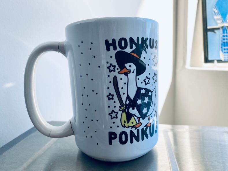Closeup of a coffee mug with a broken, then repaired, handle. On the front of a mug, a goose in a witch costume surrounded by little stars and the words HONKUS PONKUS.