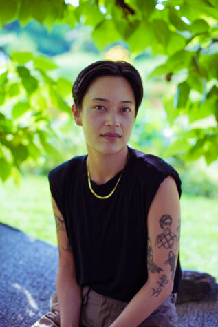 Sabrina, a person with black hair and dark eyes wearing a black sleeveless tshirt and a gold chain necklace, sitting outside under a leafy tree, looking at the camera. 