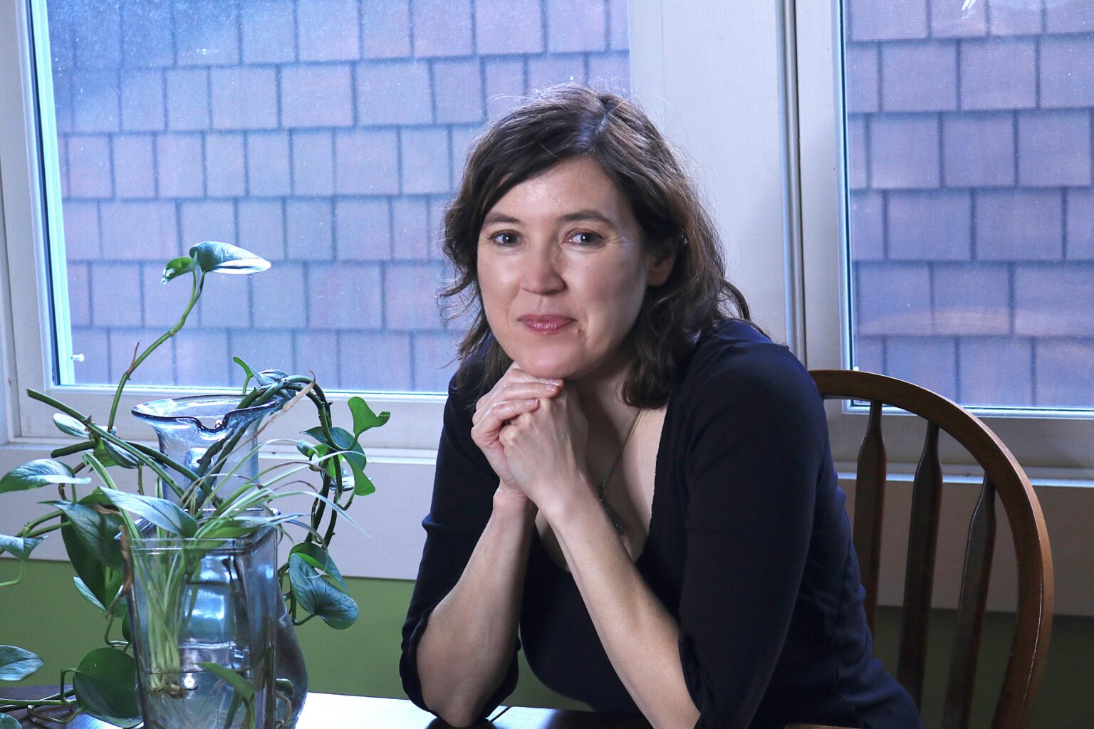 Photograph of Madeline Ostrander sitting at a kitchen table, smiling at the camera with her hands folded beneath her chin. Behind her are windows and a green wall. Beside her are plant cuttings propagating in vases of water.