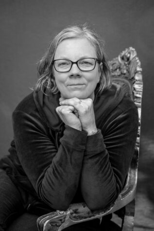 A black-and-white portrait of a white woman with glasses and gray hair sitting on a chair, her chin in her hands. She is looking at the camera and half-smiling. 