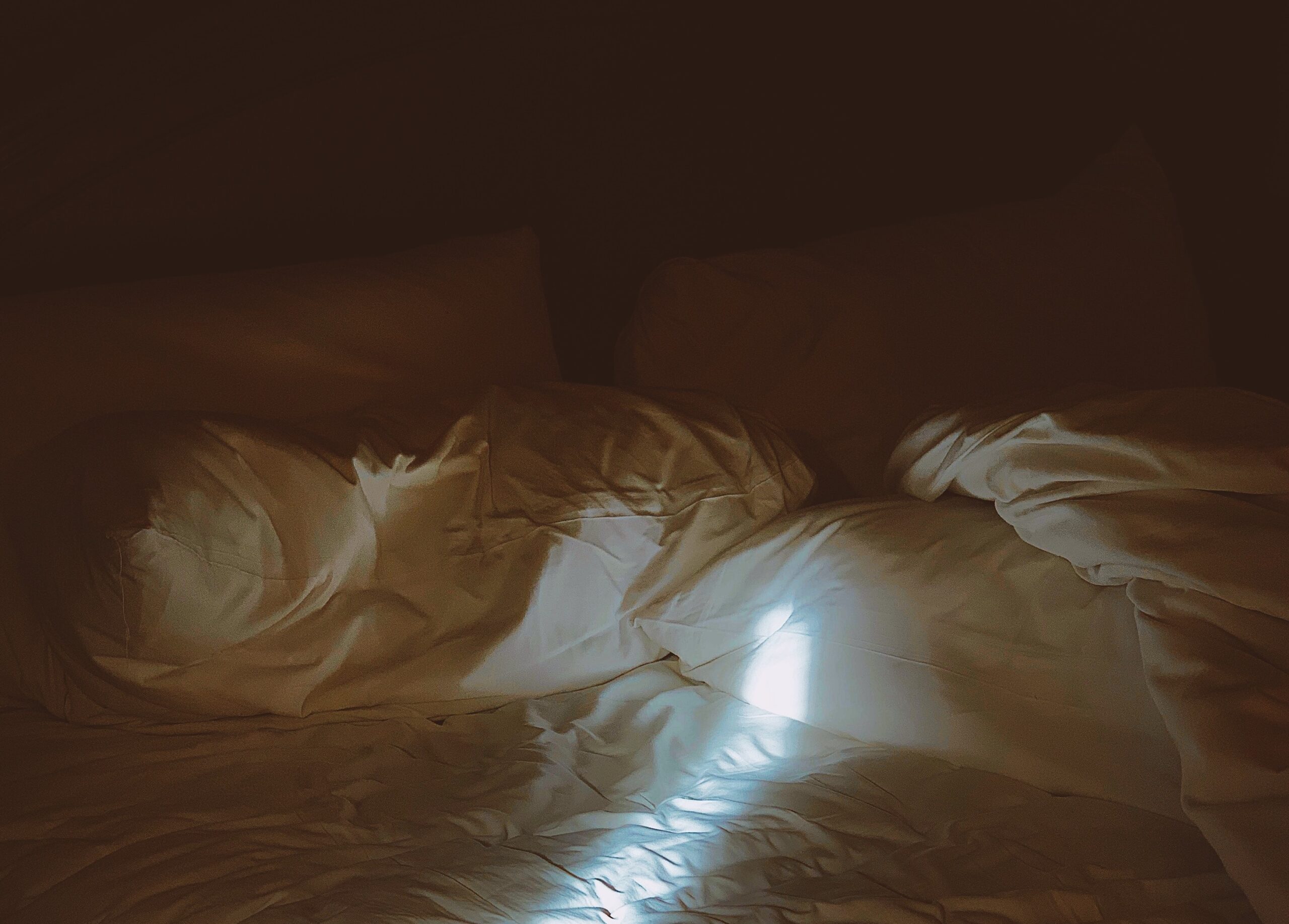 A beam of light falls on an unmade bed in a dark room.