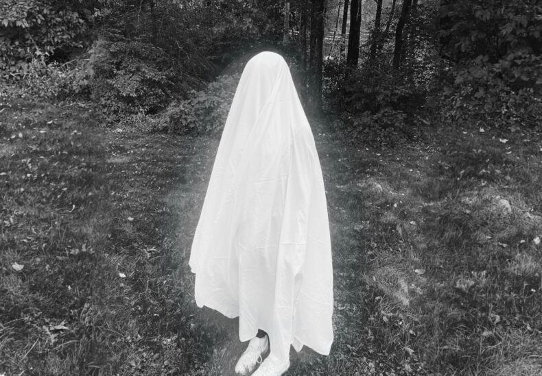 Black and white photo of a bedsheet ghost standing outside in the grass. The person is small, and their white sneakers can be seen. They are glowing faintly.