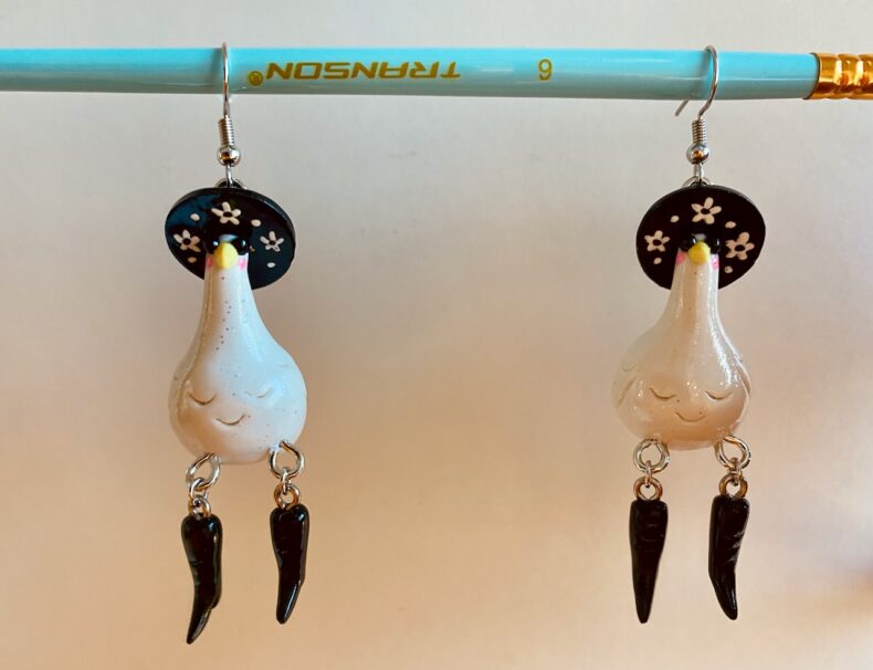 A pair of earrings dangling from a robin's-egg-blue paintbrush. The earrings are sparkly white geese wearing flowery witch hats, sunglasses, and high-heeled black boots.