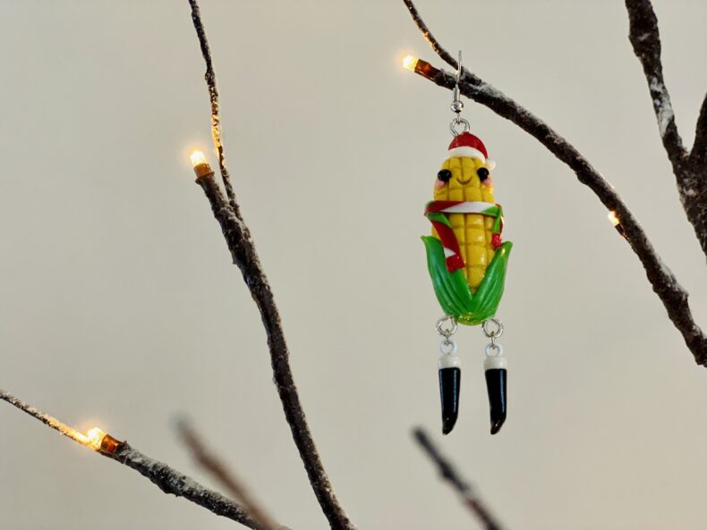 Closeup of pre-lit, artificial bare branches hung with a dangly earring shaped to look like a smiling ear of corn wearing a santa hat, a festive scarf, and kicky little black boots.