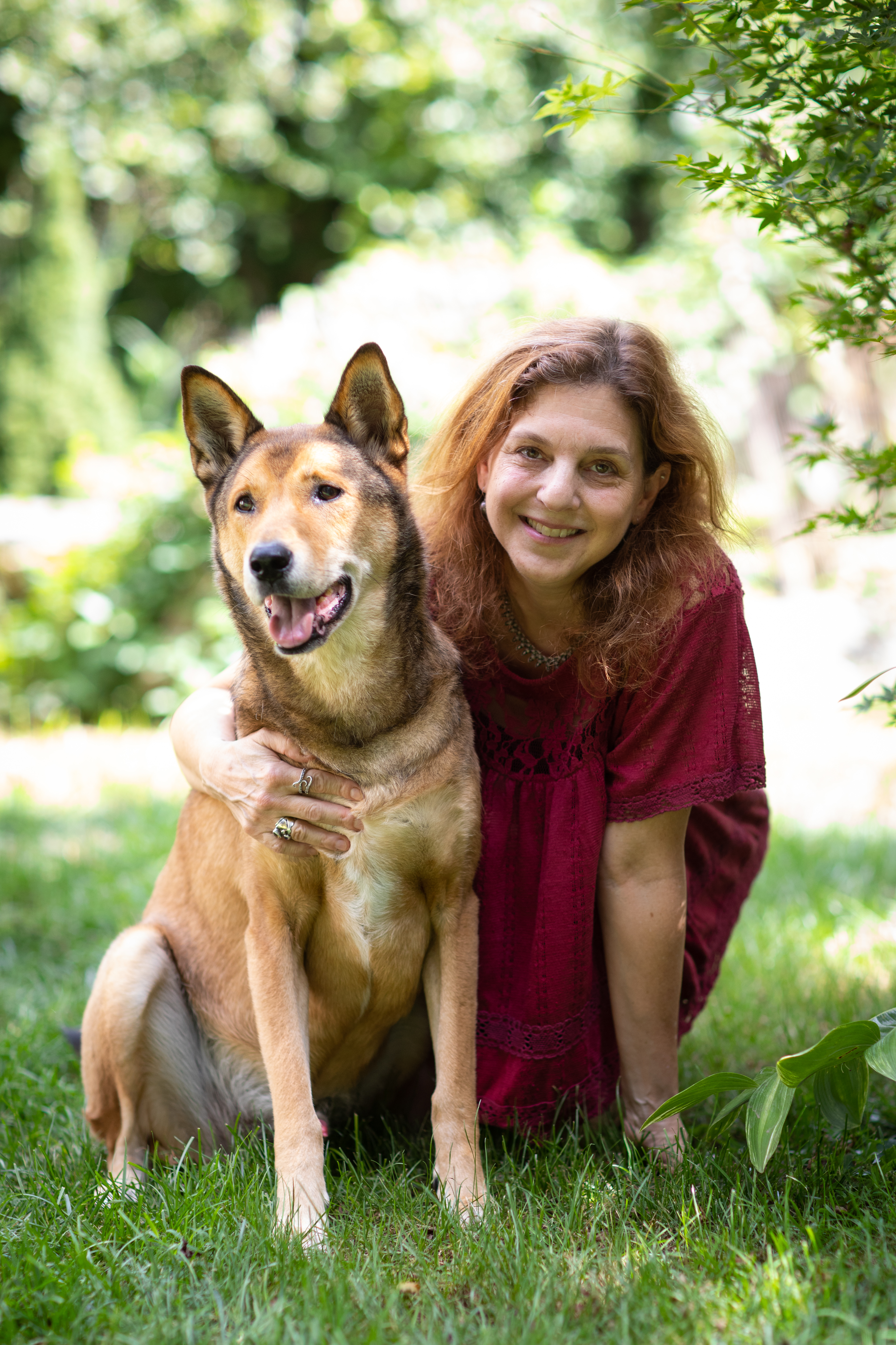 Portrait of Jennifer S. Holland sitting on the grass, hugging her dog and smiling at the camera. 
